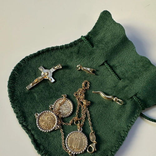 A collection of gold religious and lucky jewelry, including the Saint Benedict crucifix, arranged on a dark green velvet drawstring pouch.