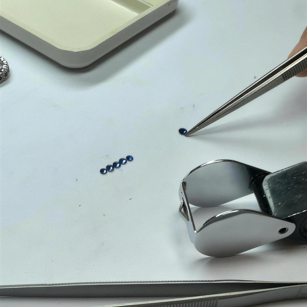 Small blue Sapphires on a white work surface with tweezers, a metal clamp tool, and a tray visible in the background. We are in a gemologist studio