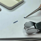 Small blue Sapphires on a white work surface with tweezers, a metal clamp tool, and a tray visible in the background. We are in a gemologist studio
