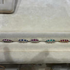 Six 18k gold rings with colorful precious gemstone clusters are displayed side-by-side on a beige velvet jewelry organizer. There are rings with Ruby, Emerald, Sapphires and Diamonds