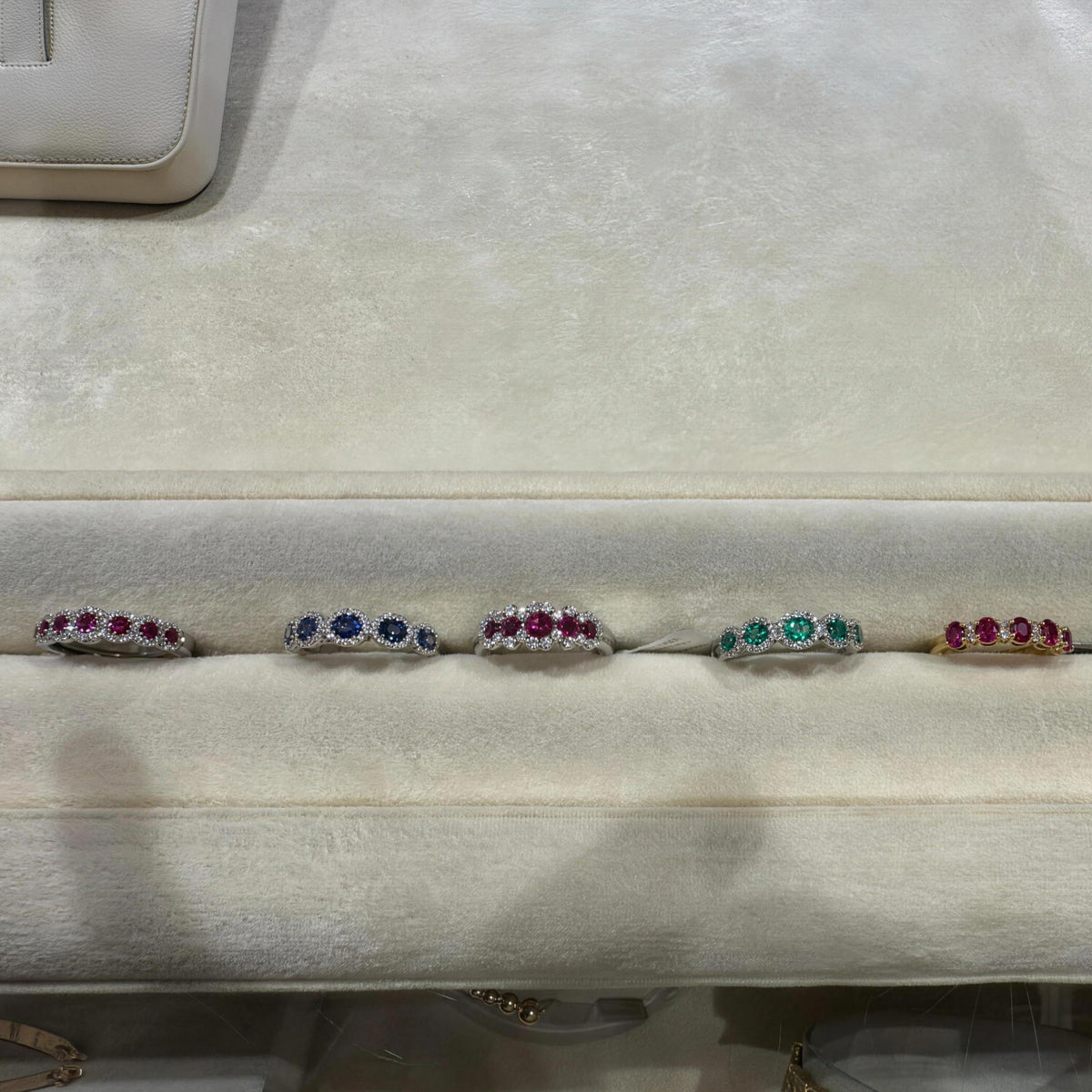Six 18k gold rings with colorful precious gemstone clusters are displayed side-by-side on a beige velvet jewelry organizer. There are rings with Ruby, Emerald, Sapphires and Diamonds