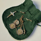 A collection of gold religious and lucky jewelry, including a crucifix and horns, as well as the 3-coin necklace, arranged on a dark green velvet drawstring pouch.