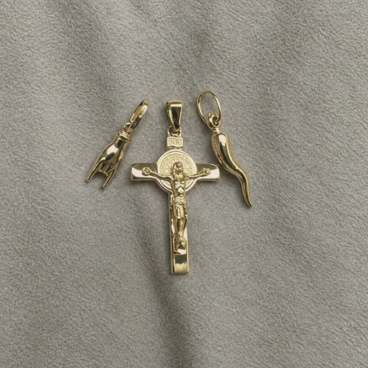 Three 18k Italian gold pendants. Crucifix,  Italian Hand & Italian Horn, arranged on a textured gray fabric background.
