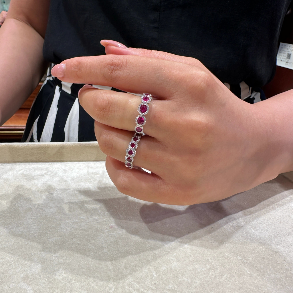 A close-up of a person's hand wearing a 18k White Gold ring set with alternating oval red and round Ruby, shown in a jewelry store setting.