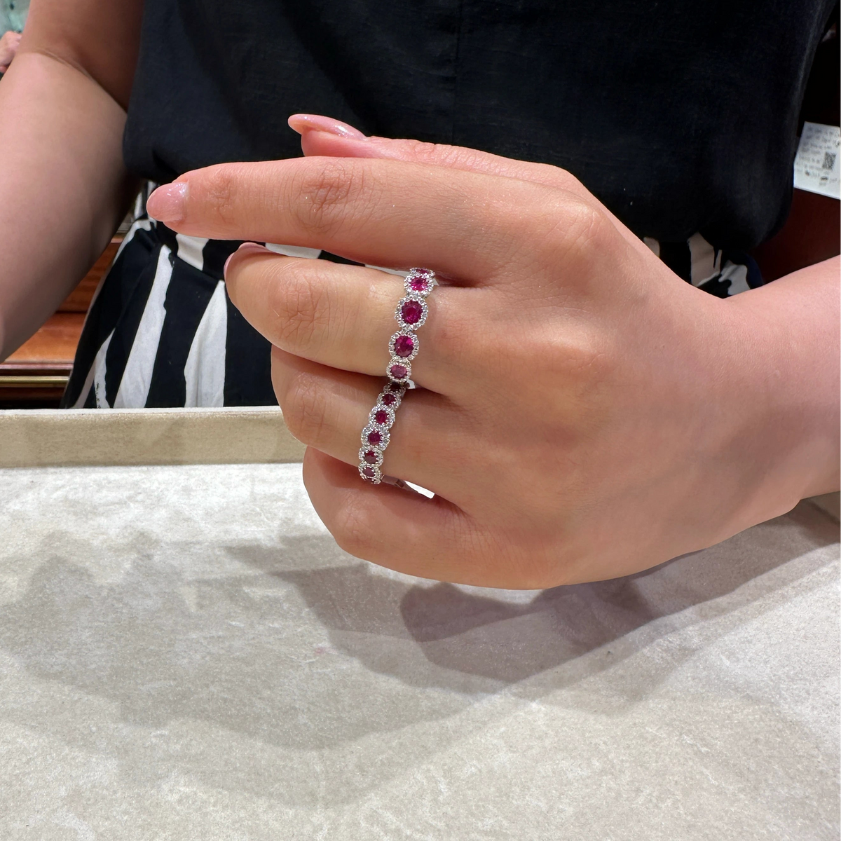 A close-up of a person's hand wearing a 18k White Gold ring set with alternating oval red and round Ruby, shown in a jewelry store setting.