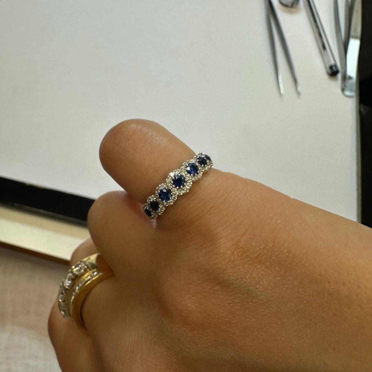 A close-up of a hand wearing the Sapphires & Diamonds ring, alongside another gold ring, with gemologist tools on a light surface in the background.