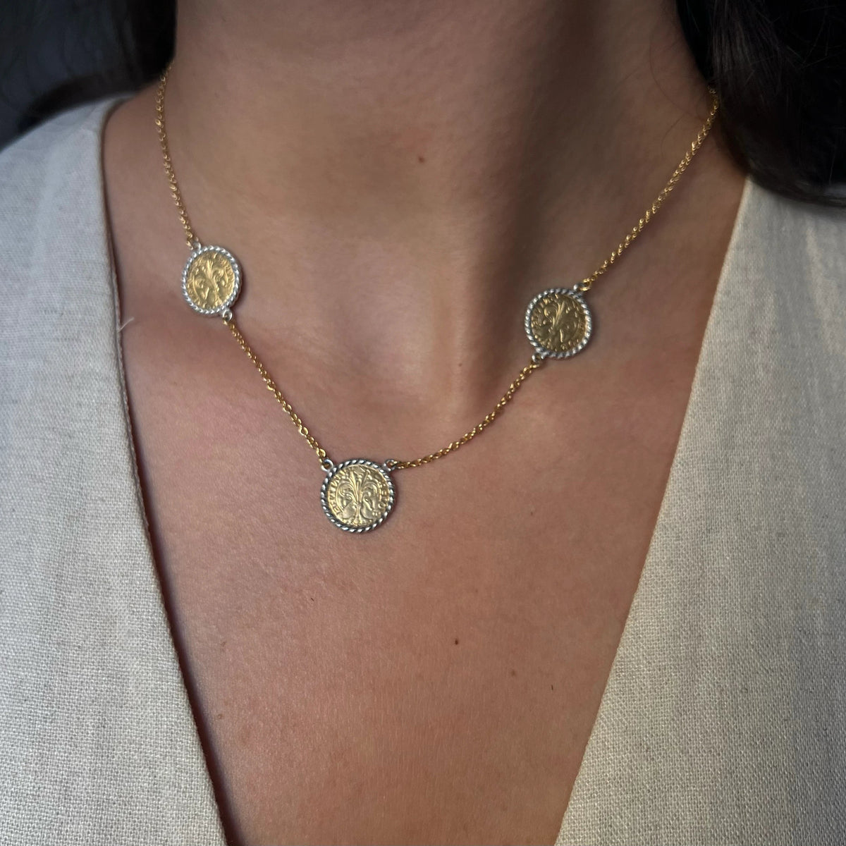 A close-up of a person wearing a delicate gold chain necklace with three round florin coin, styled with a light beige linen top.