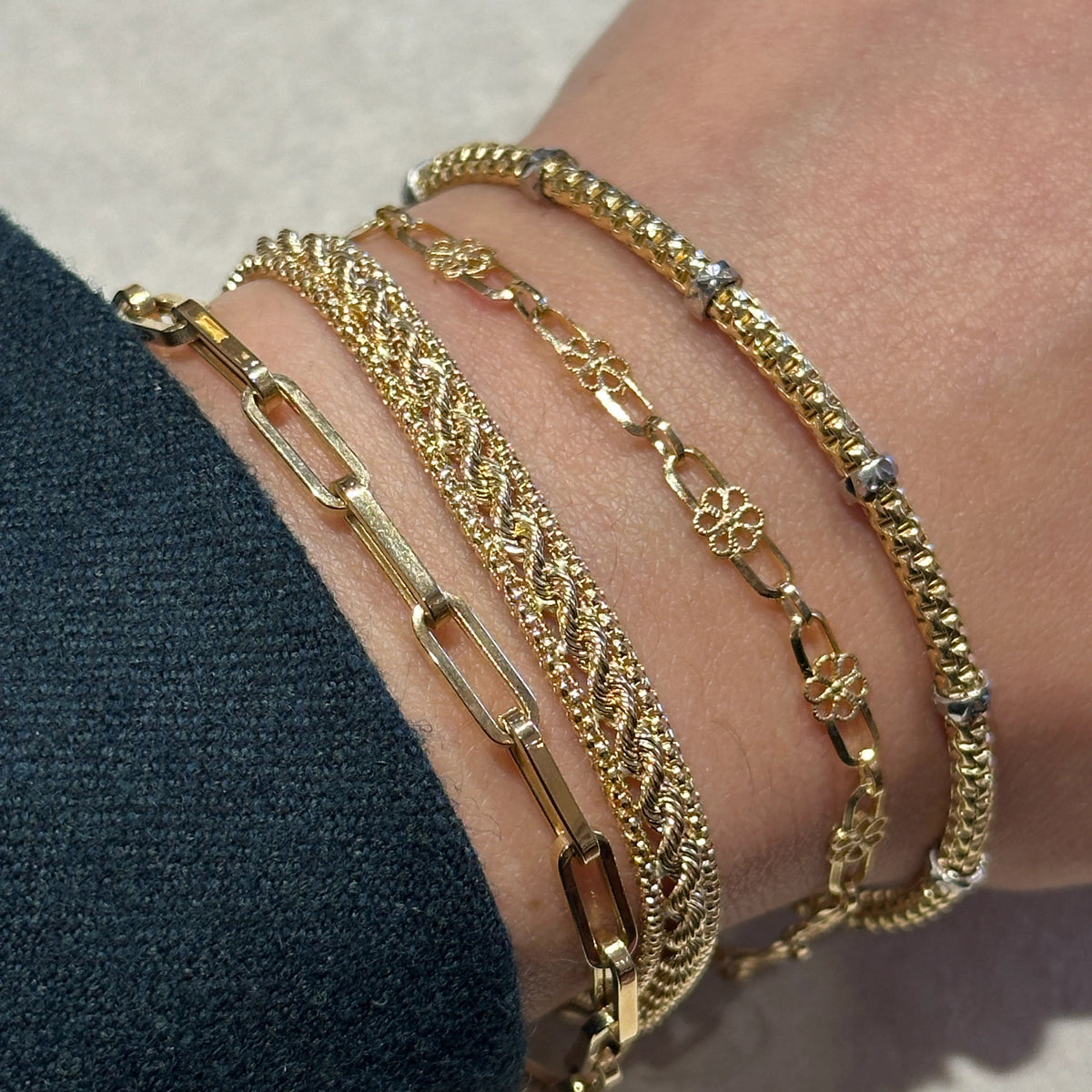 A close-up of a person's wrist wearing five stacked gold bracelets, including the floral bracelet.