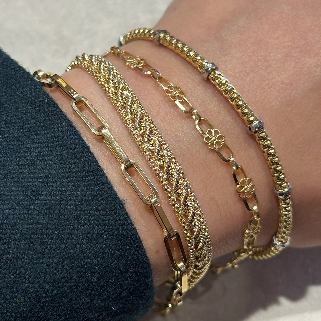 A close-up of a person's wrist wearing five stacked gold bracelets, including the paperclip bracelet.